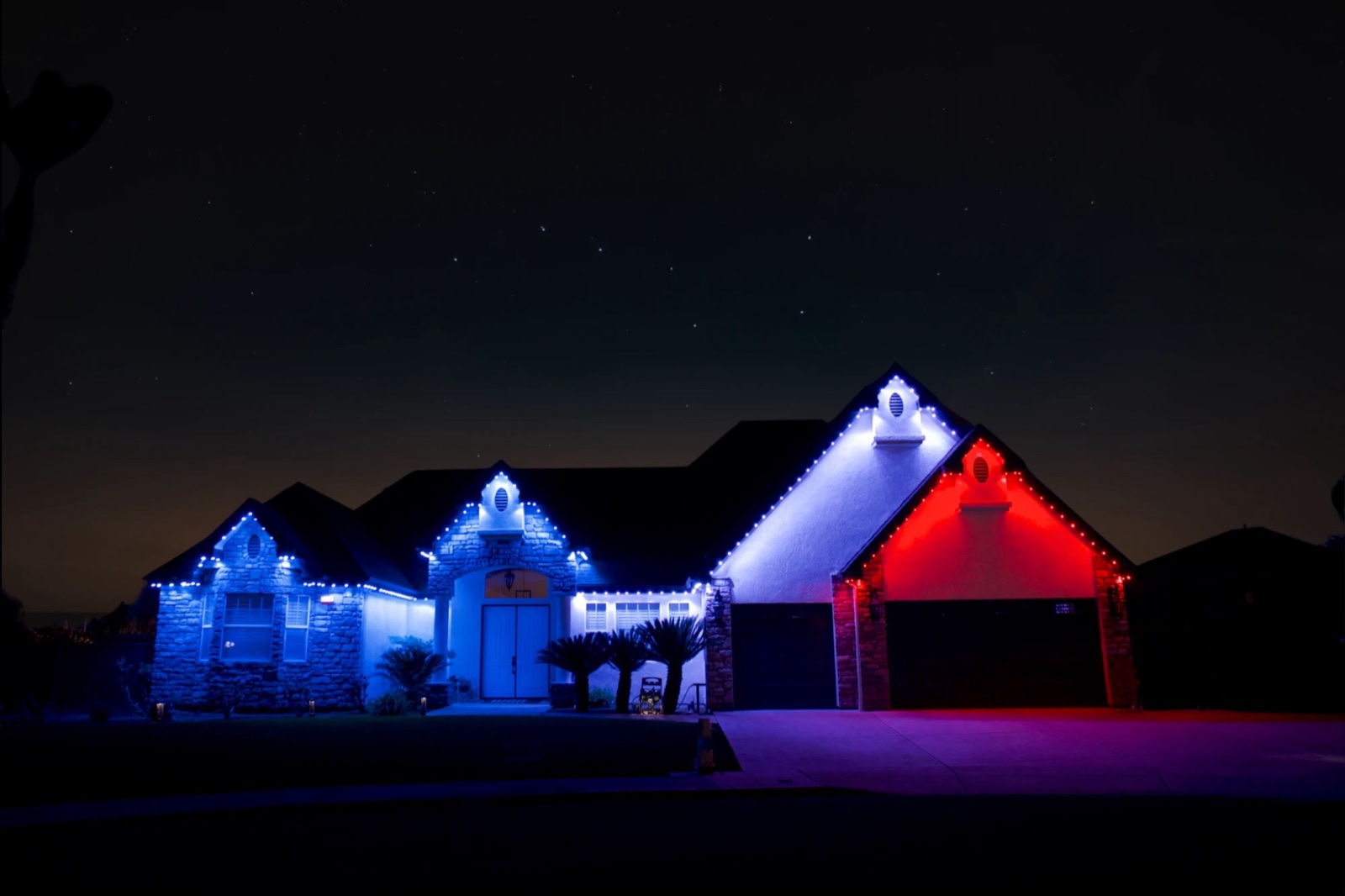 Stone and stucco home under a starry sky, rooflines lit blue and white with the garage gable in red